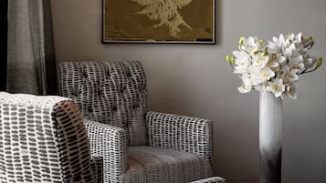 Ein Zimmer mit einem gemusterten Sessel, einem gerahmten Pflanzenprint und einer Vase mit weißen Blumen.