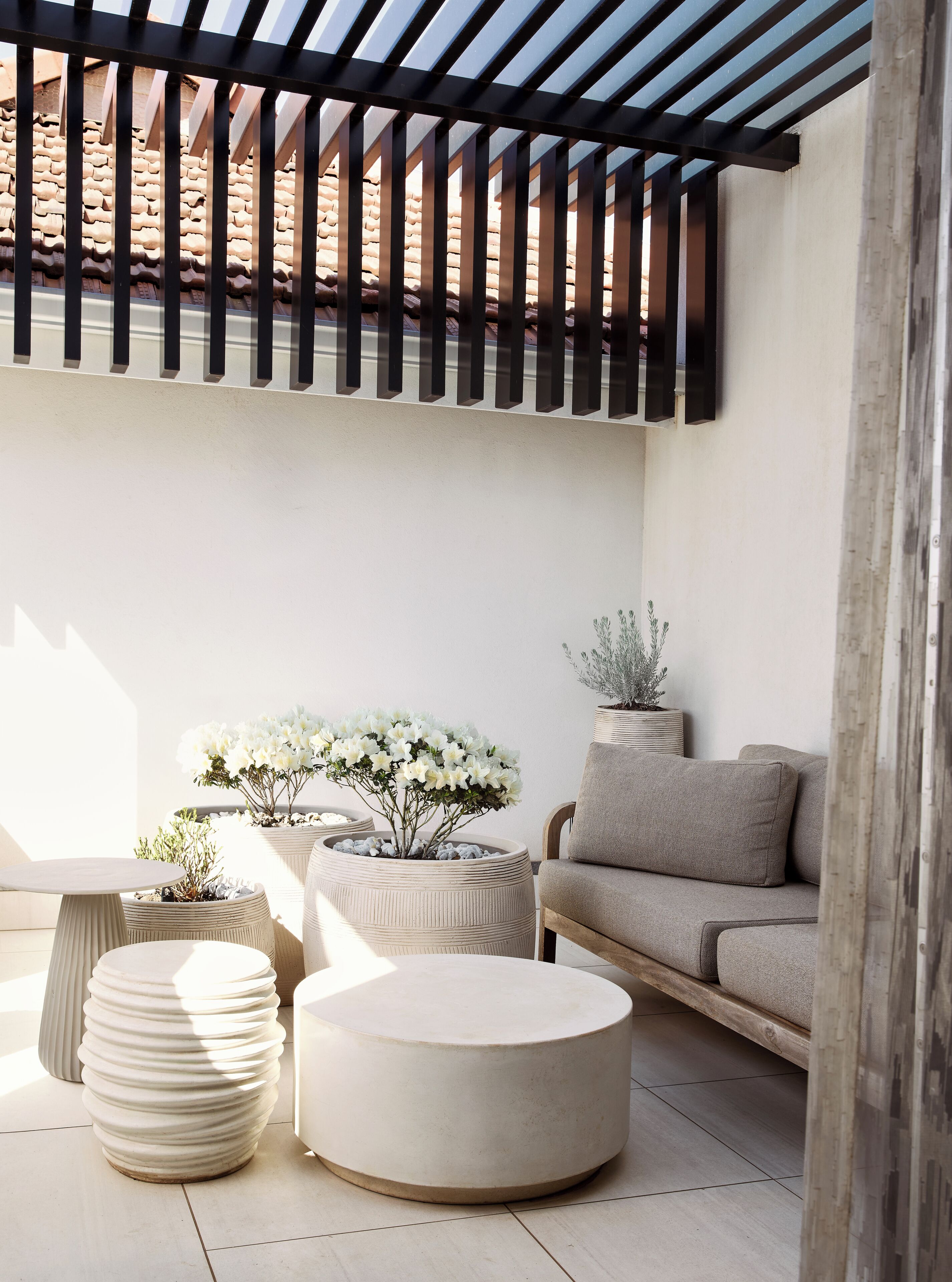 A sunlit patio with a wooden pergola, a beige sofa, round planters with white flowers, and a tiled floor.