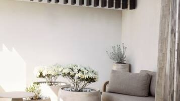 A sunlit patio with a wooden pergola, a beige sofa, round planters with white flowers, and a tiled floor.