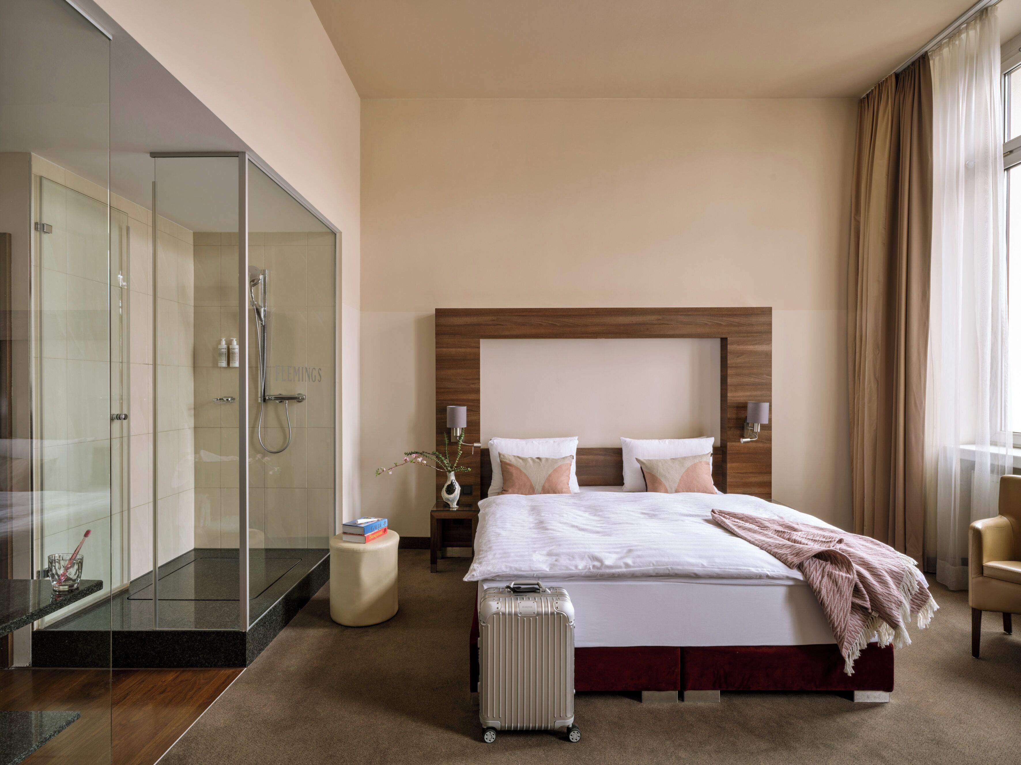 Una habitación de hotel con una cama grande, una mesita de noche, una ducha con cerramiento de vidrio y una maleta en el suelo.