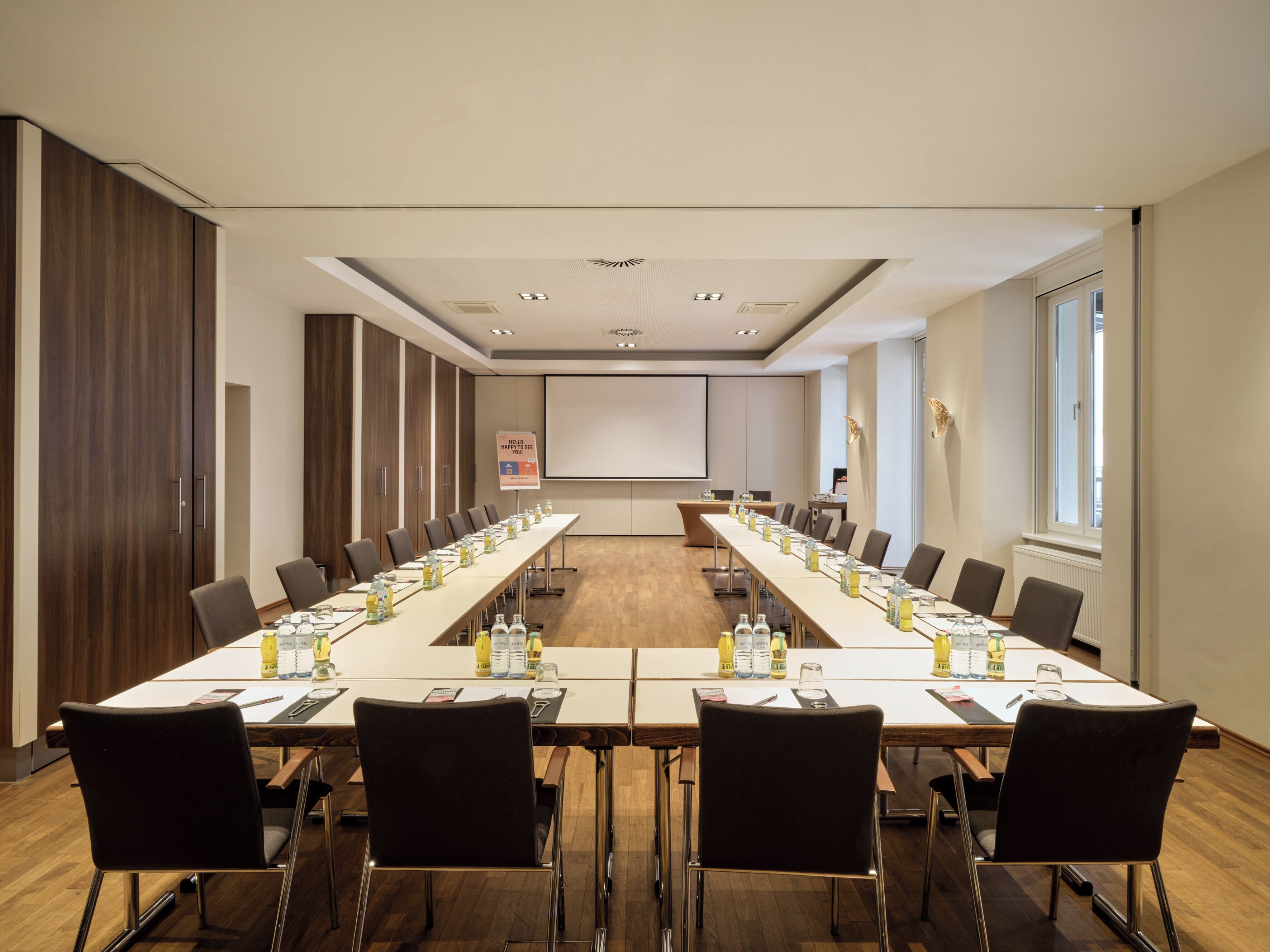 A conference room with a long table, chairs, a projector screen, and a sign.
