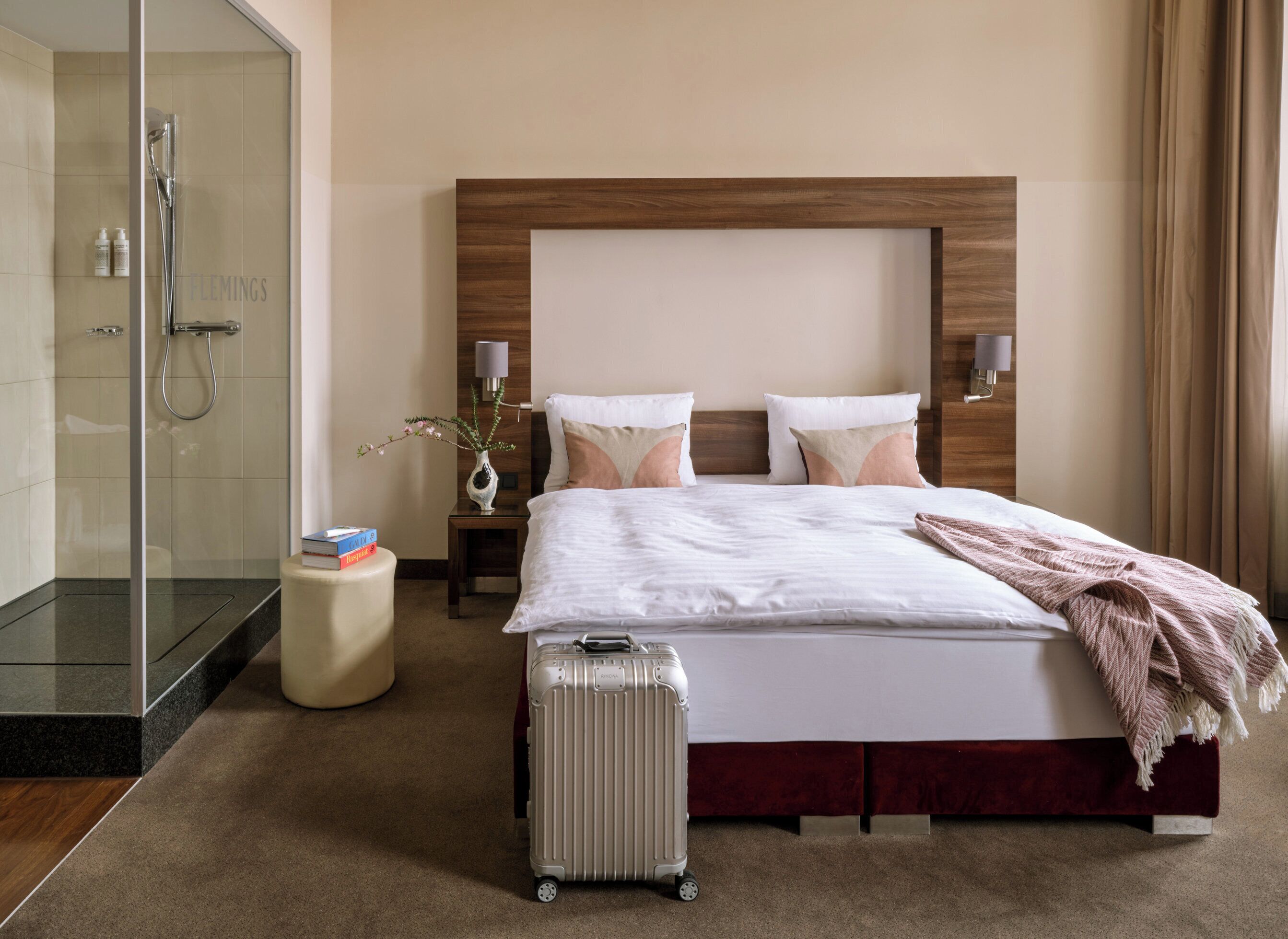 A hotel room with a bed, a bedside table, a lamp, a shower, and a suitcase.