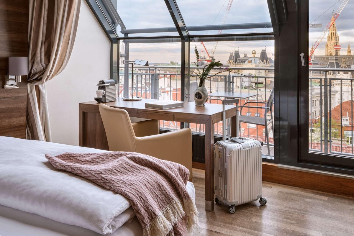 Habitación de hotel con un ventanal grande, una cama, un escritorio y vistas a la ciudad.