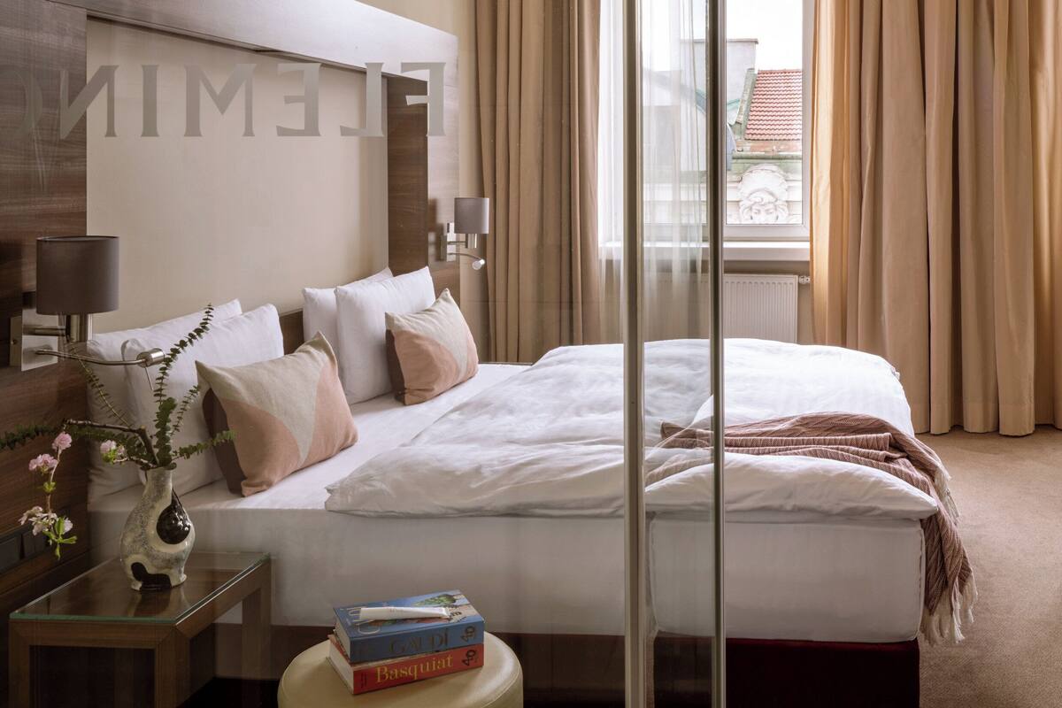 A hotel room with a large bed, a bedside table with a lamp, a vase with flowers, and a window with curtains.
