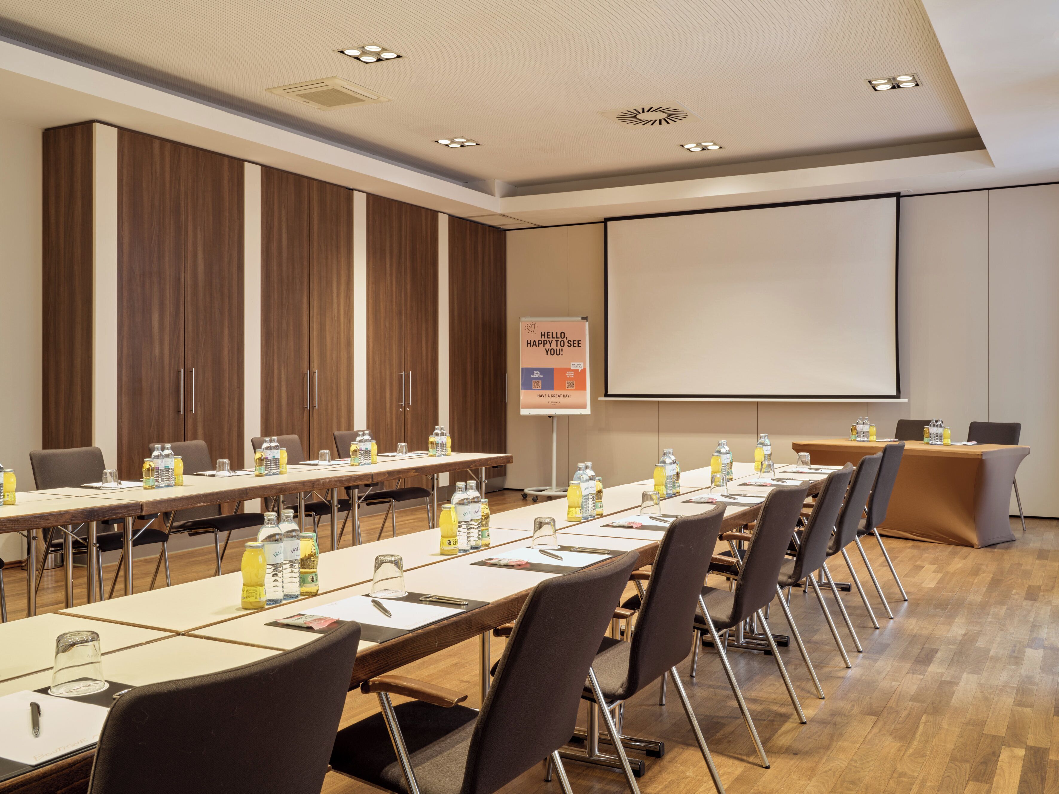 A conference room with a long table, chairs, a projector screen, and a sign with a greeting message.