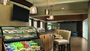 Un café moderno con un mostrador que exhibe pasteles y un refrigerador para bebidas. Hay taburetes de bar y un televisor montado en la pared.