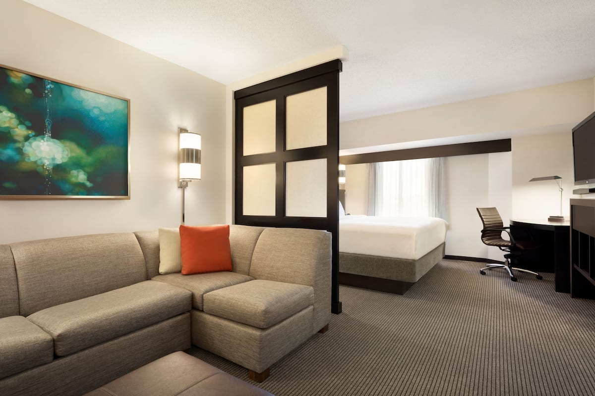 A hotel room with a sectional sofa, a bed, a desk, and a large painting on the wall.