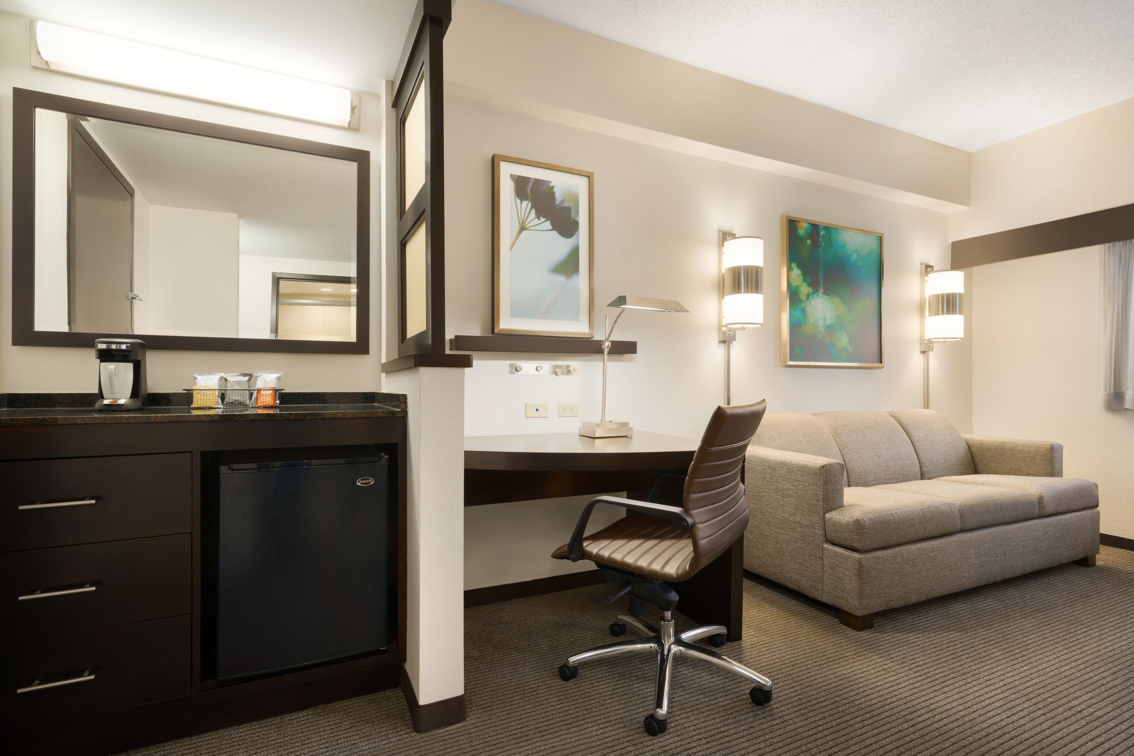 Habitación de hotel con escritorio, silla, sofá, minibar, cafetera y dos lámparas de pared.