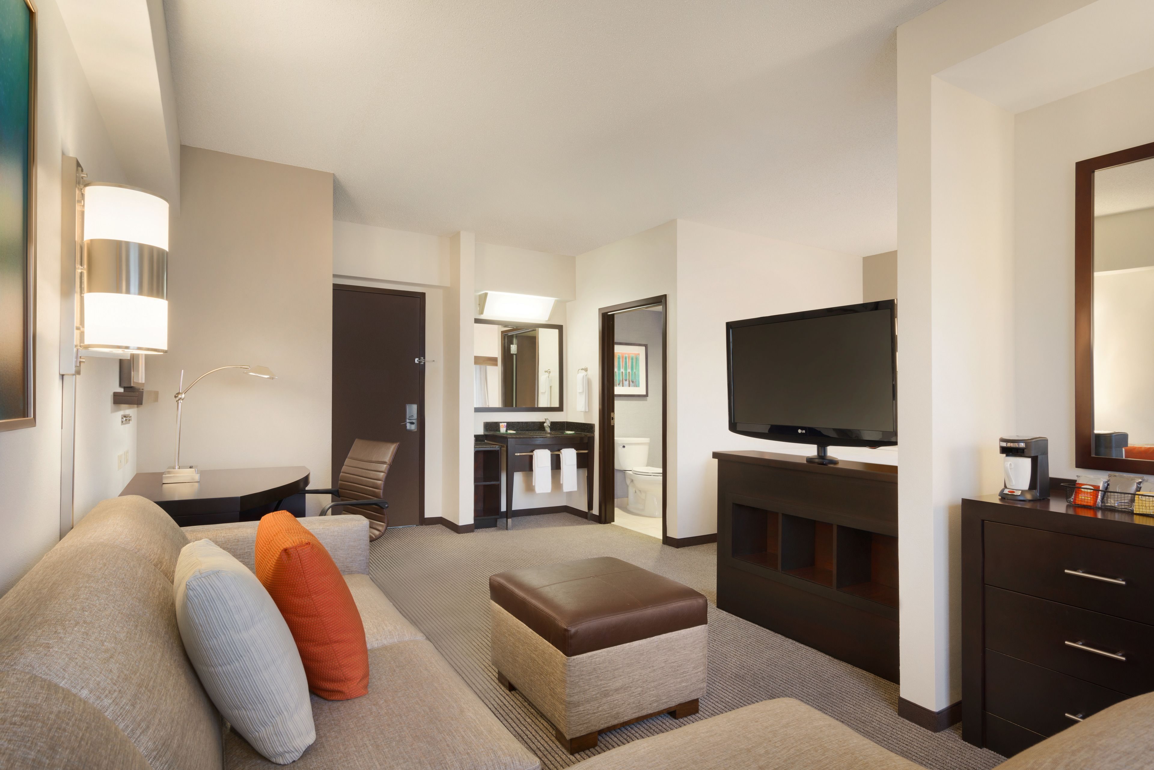 Quarto de hotel com sofá, apoio para os pés, televisão e escrivaninha com abajur.