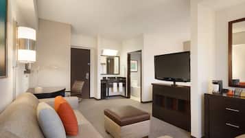 Quarto de hotel com sofá, apoio para os pés, televisão e escrivaninha com abajur.