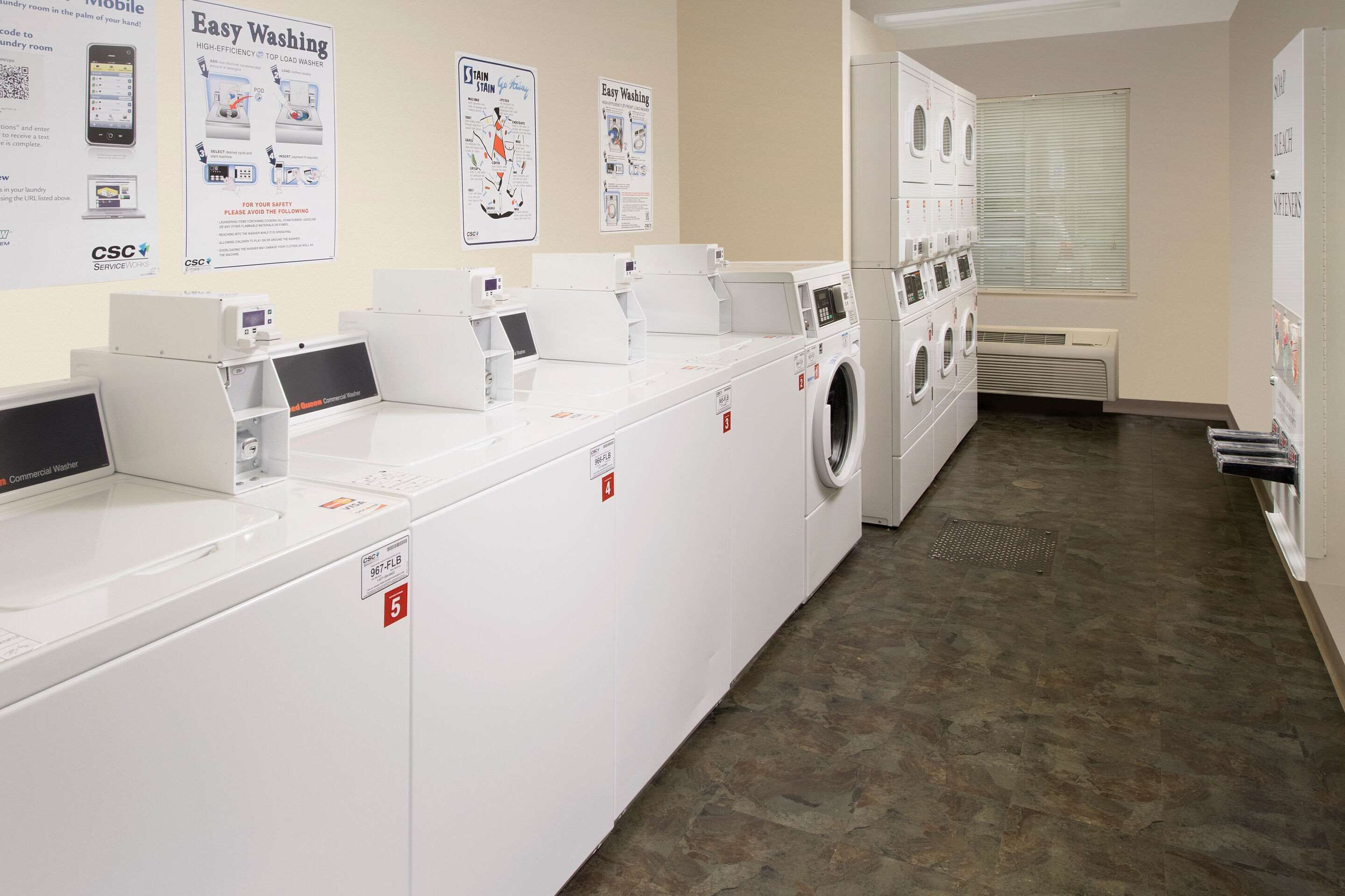 A laundry facility with multiple washing machines and dryers, instructional posters on the wall, and a tiled floor.