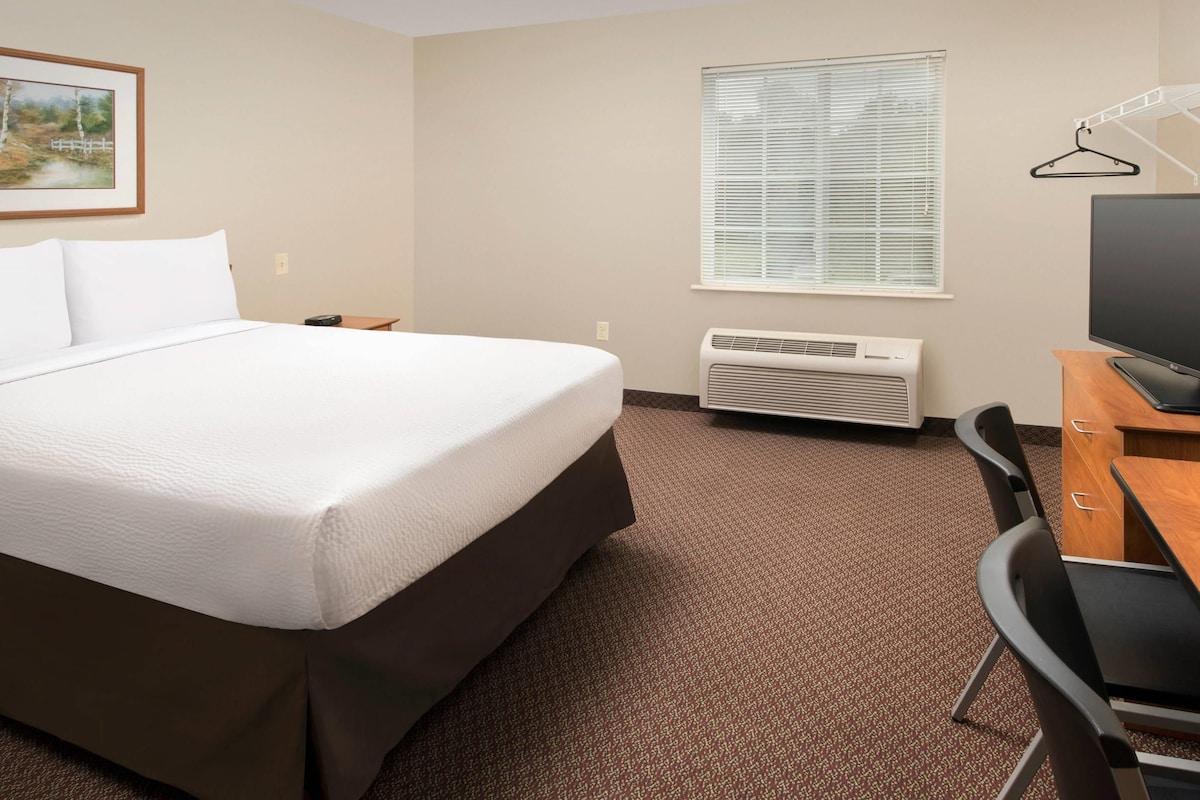 A hotel room with a bed, a desk with a TV, a window with blinds, and an air conditioning unit.