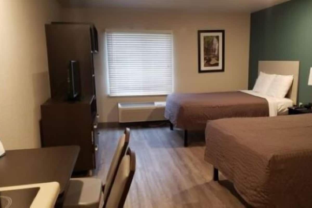 Une chambre d’hôtel avec deux lits, un bureau, une chaise, une télévision et une fenêtre avec des stores.
