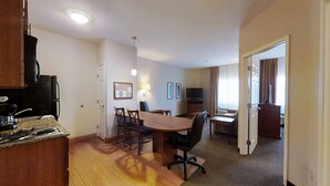 A compact hotel room with a kitchenette, a dining area, a TV, and a large window with curtains.
