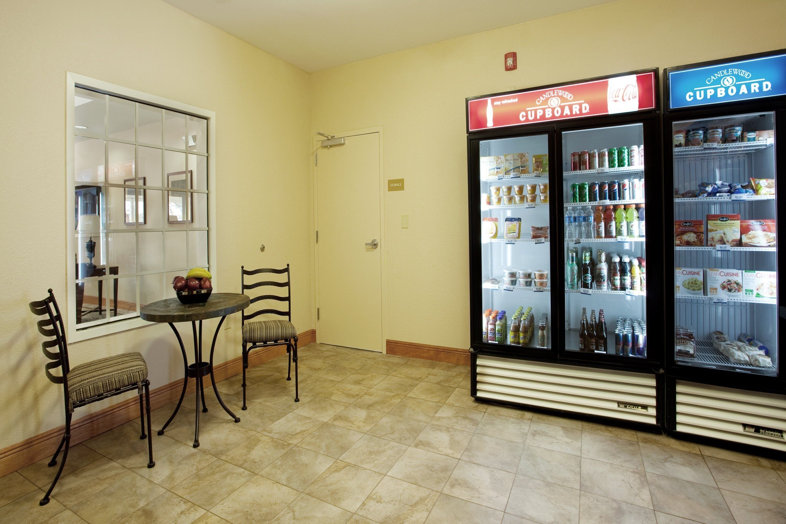 A room with a table and two chairs, a window, and two beverage vending machines labeled 'Candlewood Cupboard'.