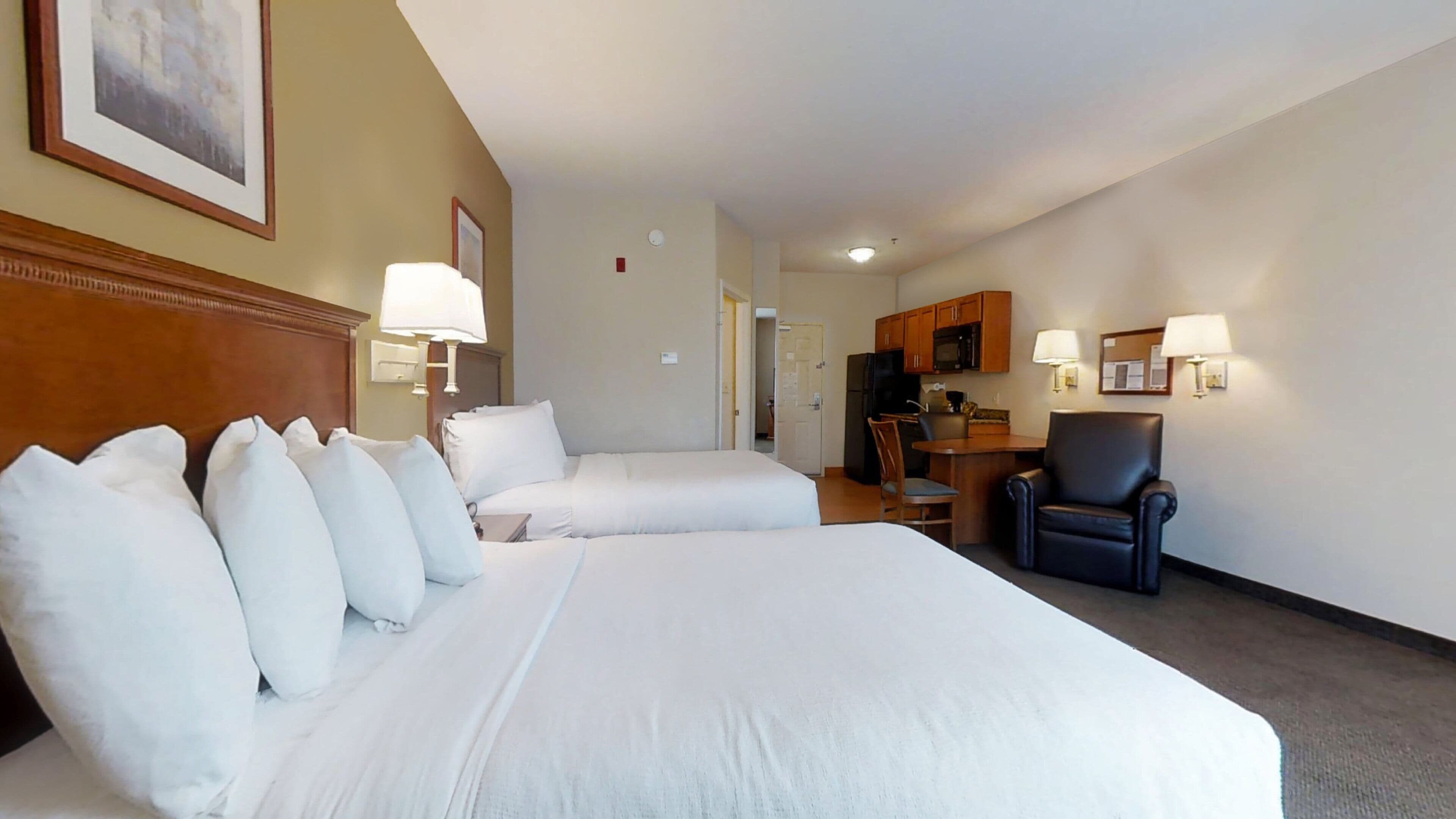 A hotel room with two beds, a desk, a chair, and a kitchenette.