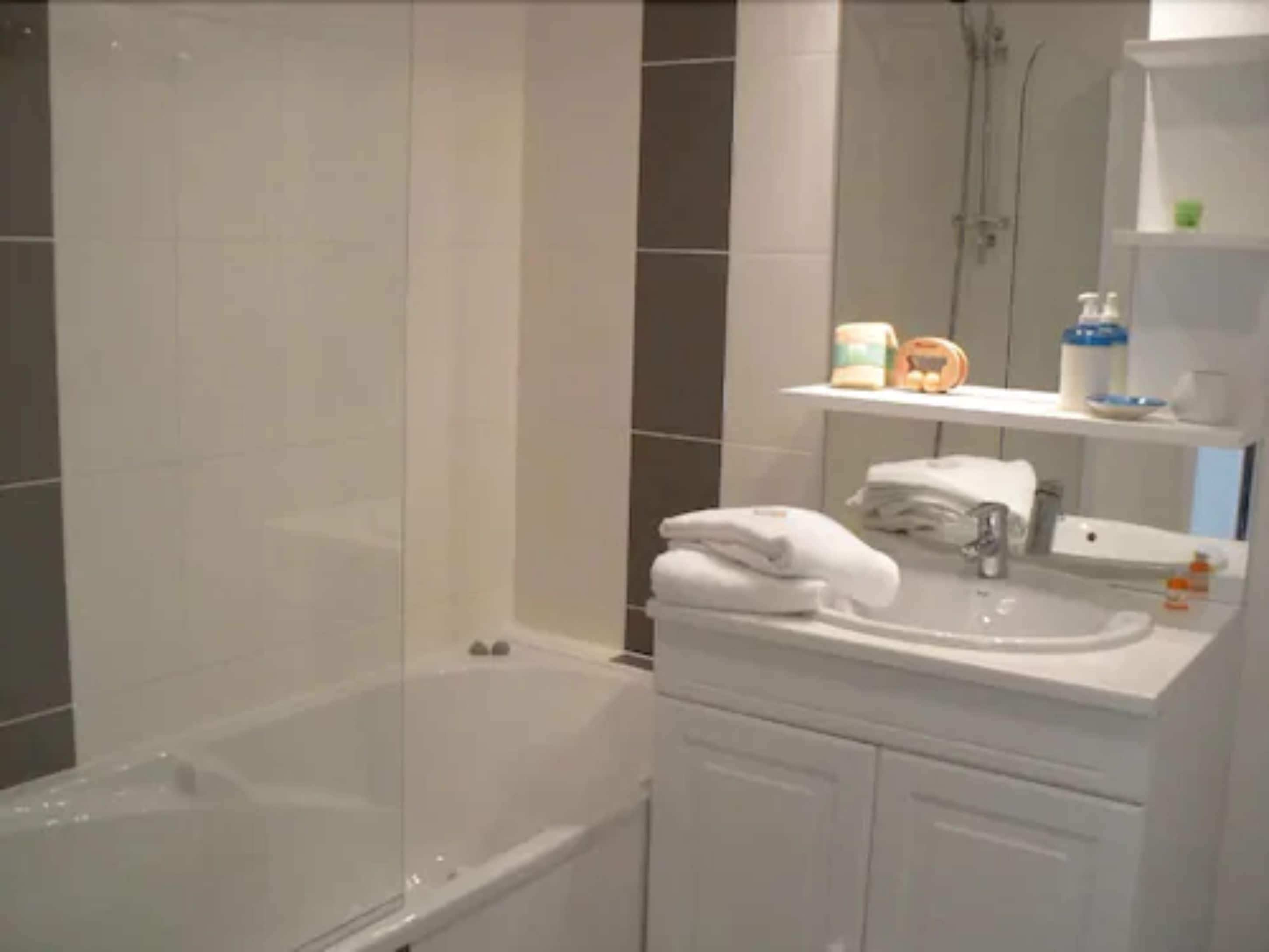 Une salle de bain avec une baignoire, un lavabo et un miroir. Des serviettes sont pliées sur le lavabo et des articles de toilette sont rangés sur les étagères.