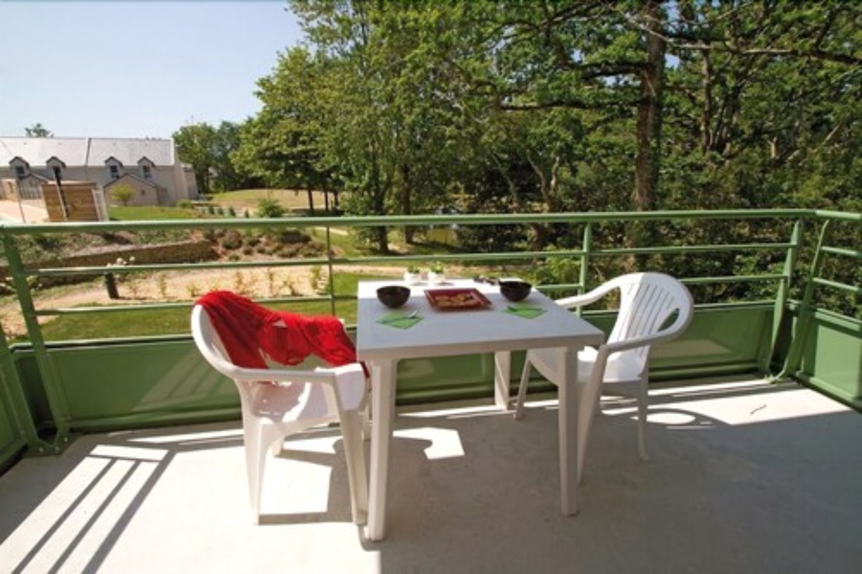 A balcony with a table and two chairs, overlooking a green landscape with trees and a building in the background.
