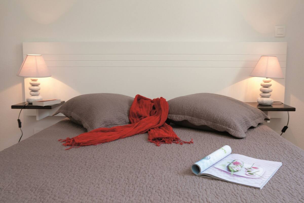 Una cama bien hecha con dos almohadas, una bufanda roja y una revista encima. Hay dos mesitas de noche con lámparas y un enchufe.