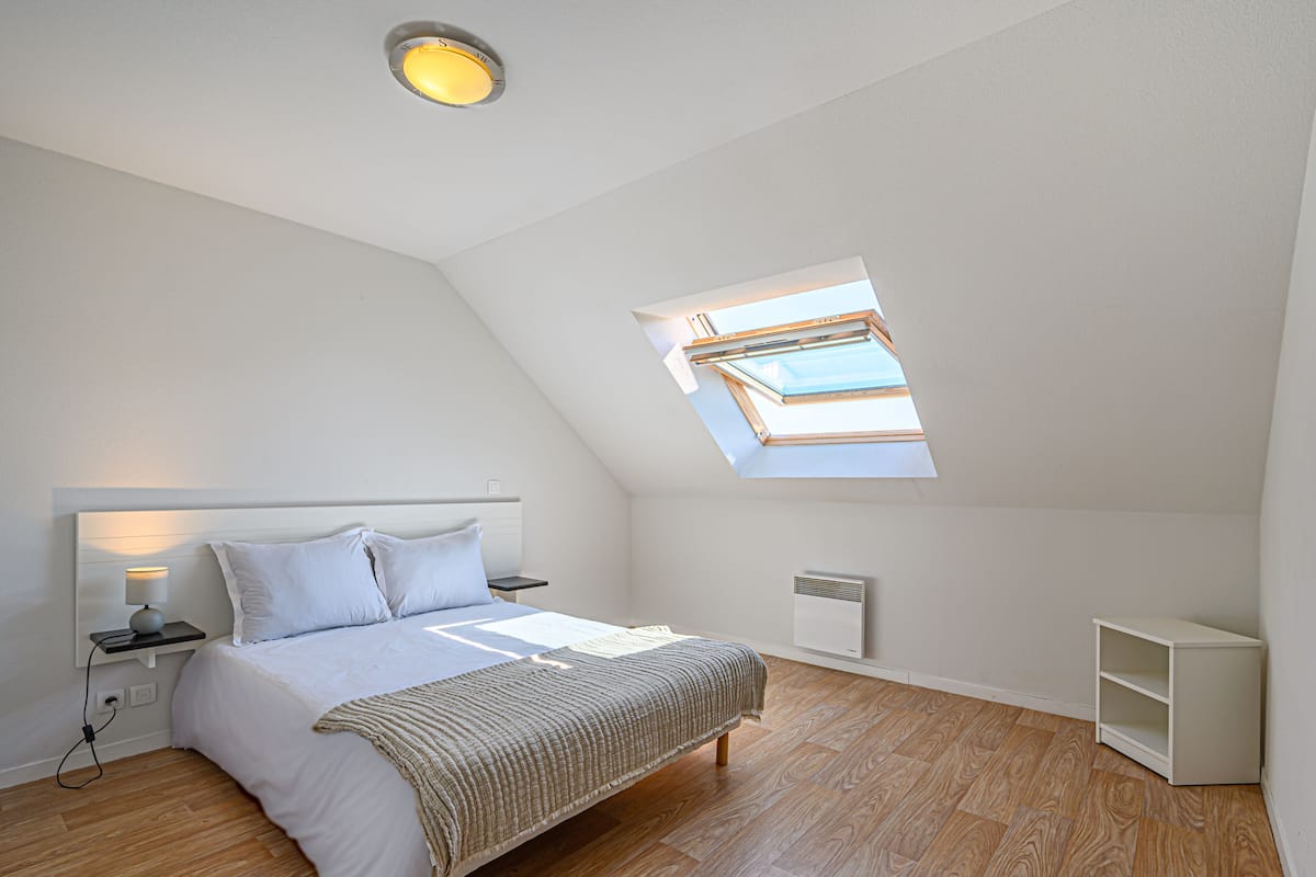 Un dormitorio con techo inclinado, tragaluz, cama con cabecera, mesitas de noche y un armario blanco.