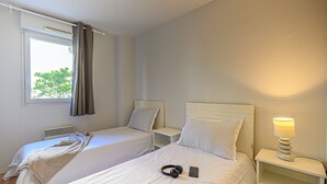 Ein Hotelzimmer mit zwei Einzelbetten, einem Nachttisch mit Lampe, einem Fenster mit Vorhängen und Blick auf Grünfläche.