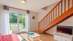 Ein Wohnzimmer mit einer Holztreppe, einem Fernseher auf einem weißen Schrank, einem roten Sofa, einem kleinen weißen Tisch und einer Glastür, die zu einem Außenbereich mit Stühlen führt.