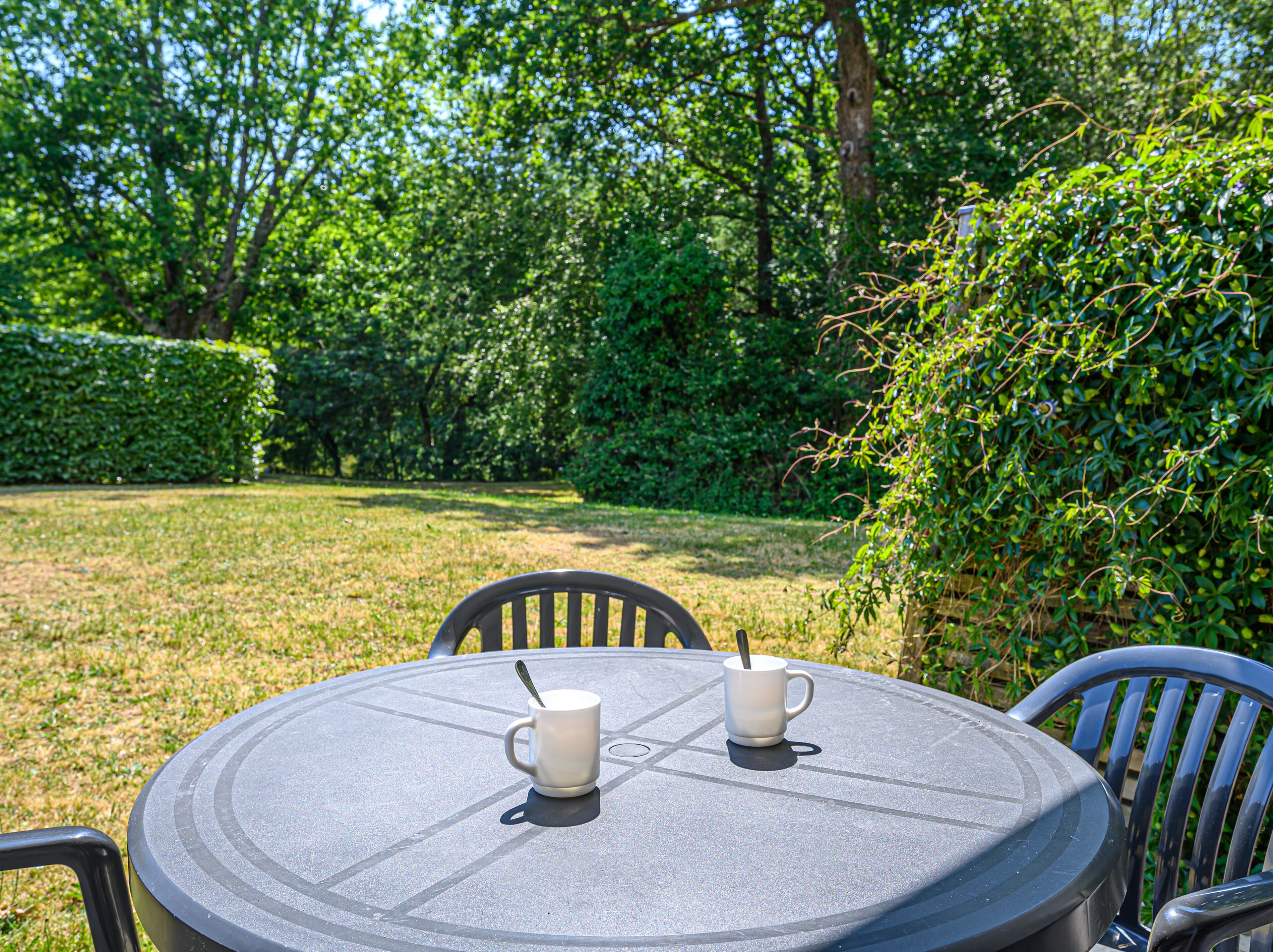 A round table with two white cups and a spoon, surrounded by chairs, set in a garden with green grass and trees.