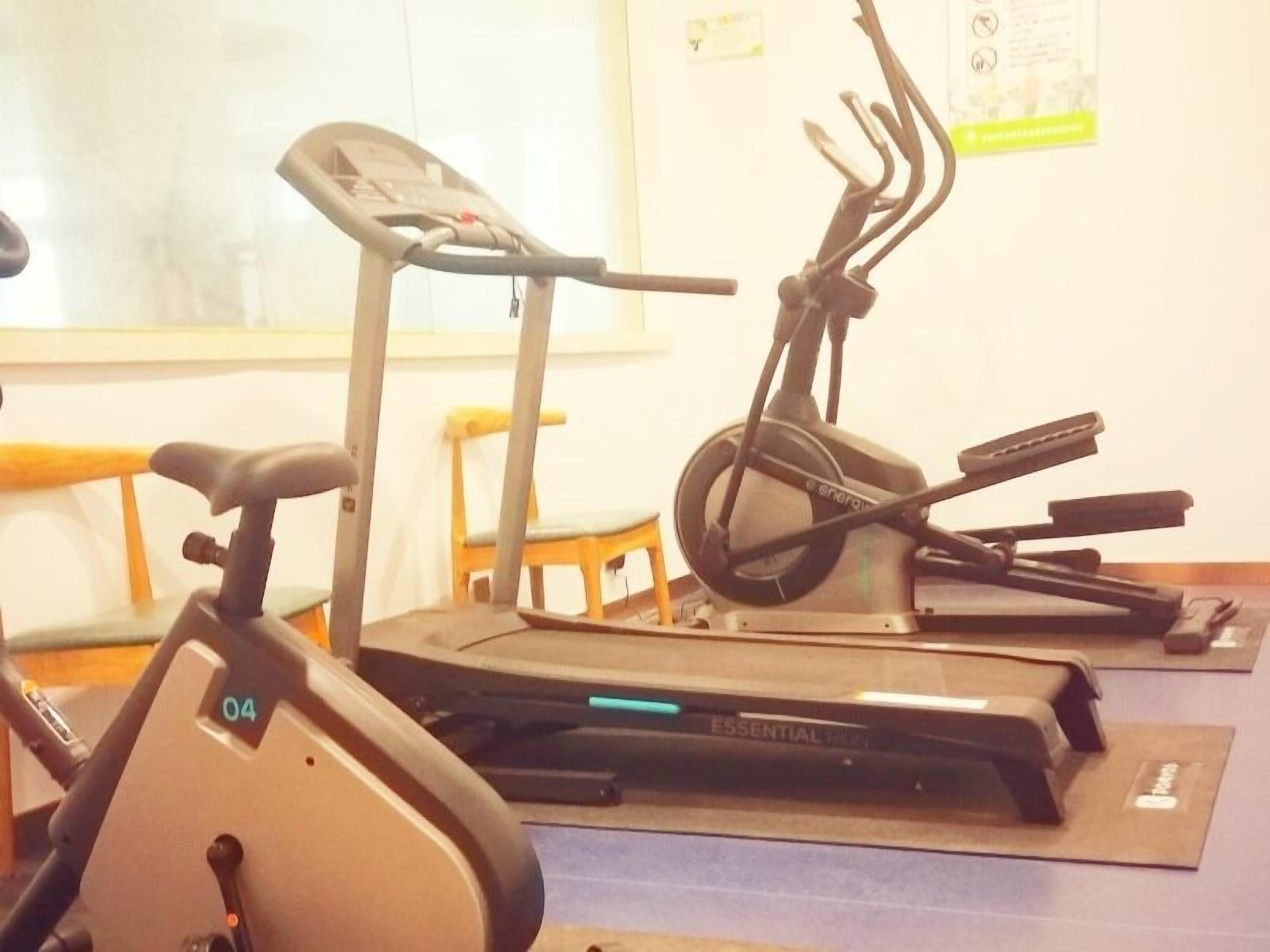 A stationary bike and a treadmill in a room with a wooden chair and a mat on the floor.