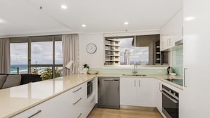 A modern kitchen with white cabinets, a stainless steel dishwasher, and an oven. There is a window with a view of buildings and a clock on the wall. - Sunbird Beach Resort (Main Beach)