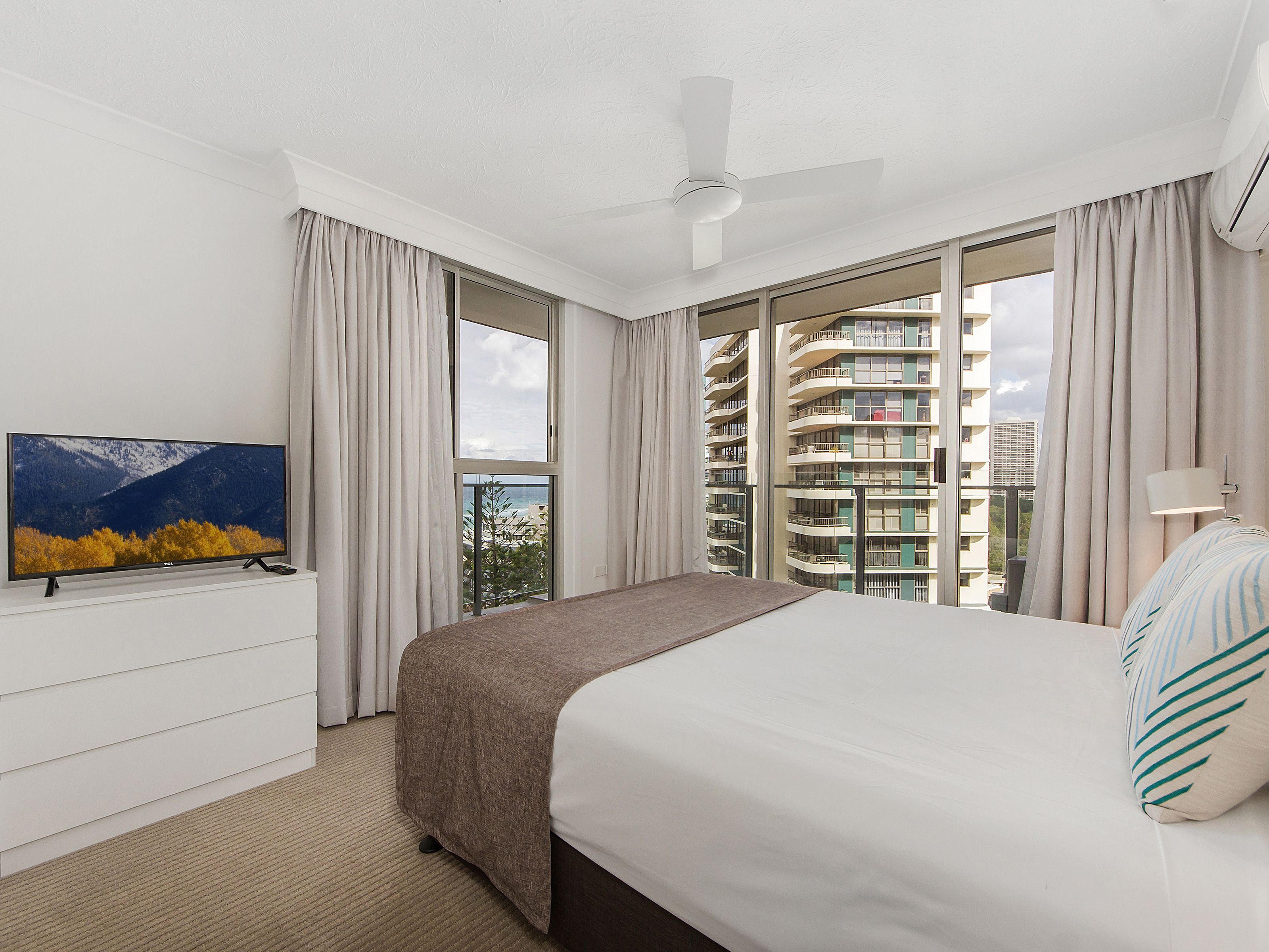 A hotel room with a bed, a TV, a ceiling fan, and large windows with curtains.