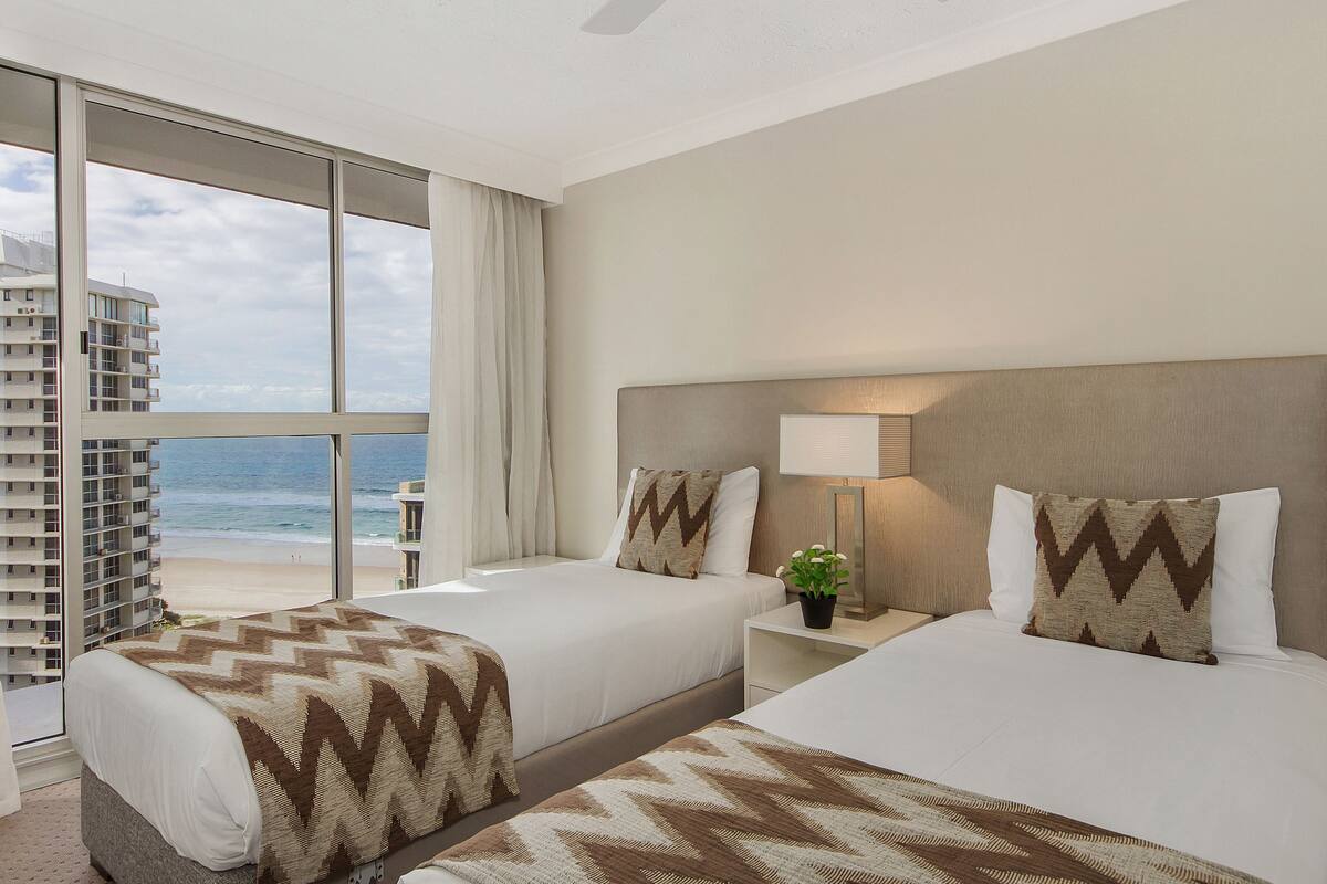 Une chambre d’hôtel avec deux lits, une grande fenêtre donnant sur la plage et l’océan, et un ventilateur de plafond.