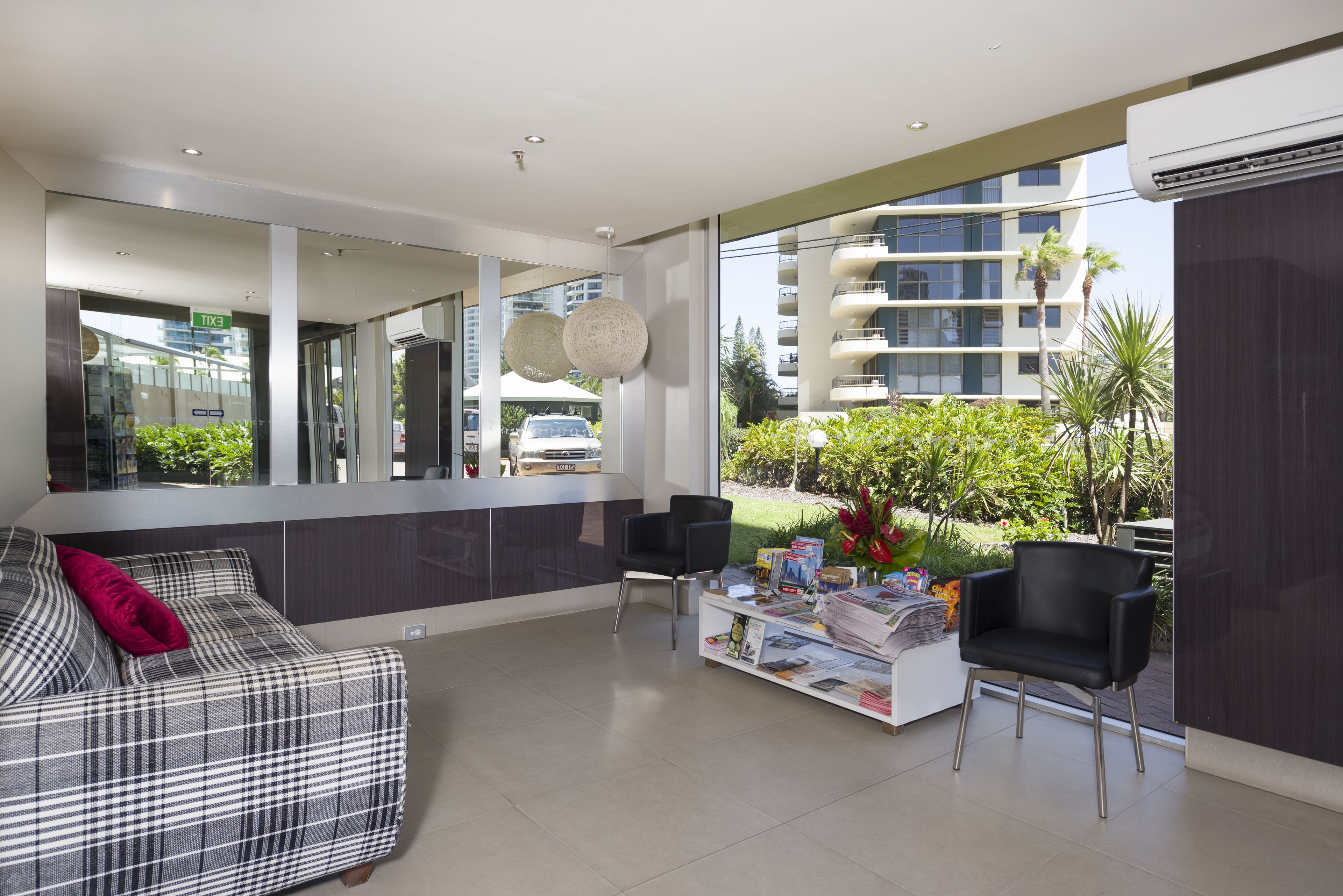 Uma sala de estar moderna com sofá, mesa de centro e cadeiras, grandes janelas com vista para um edifício e vegetação.