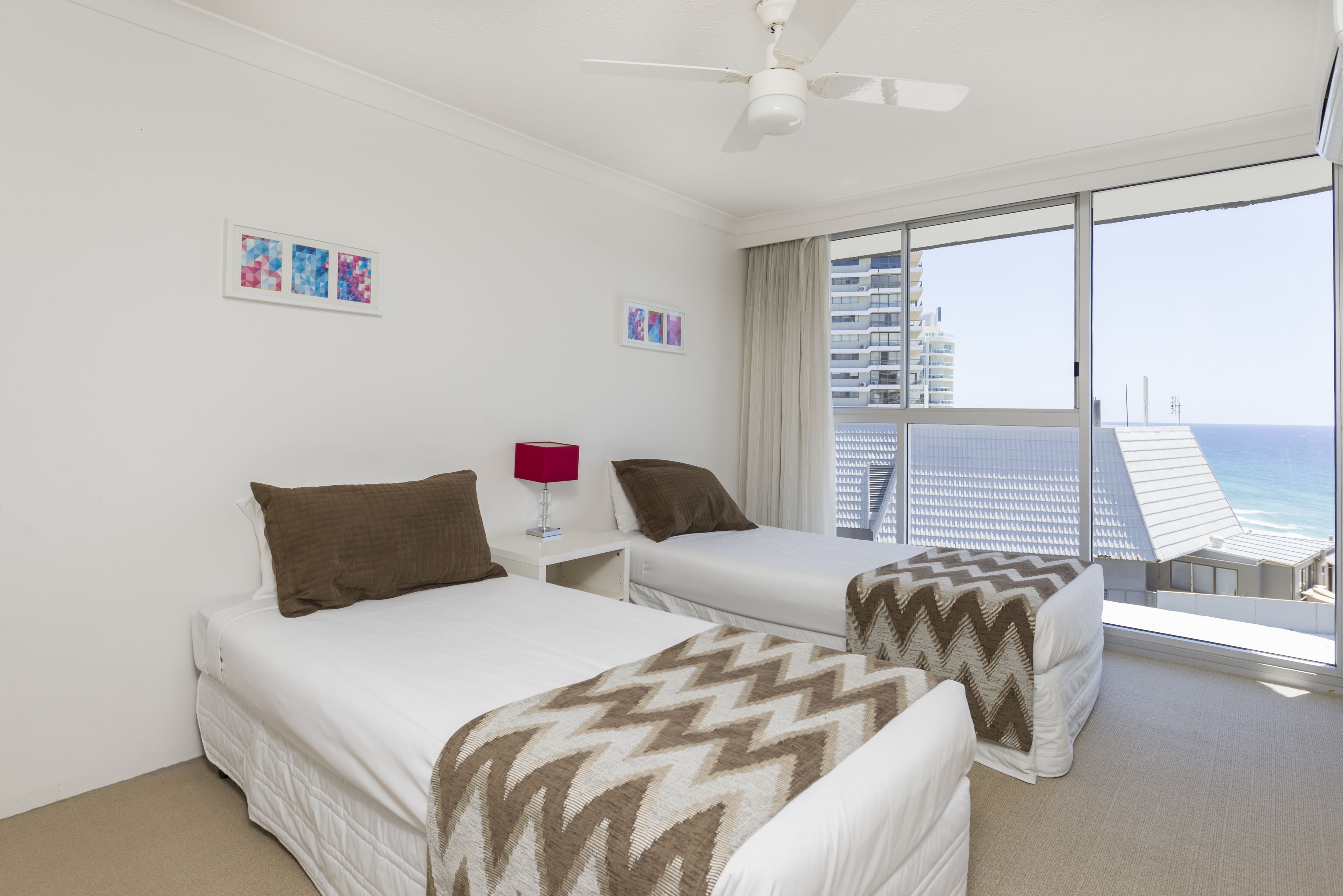 A hotel room with two beds, a ceiling fan, and a large window with a view of the ocean and buildings.