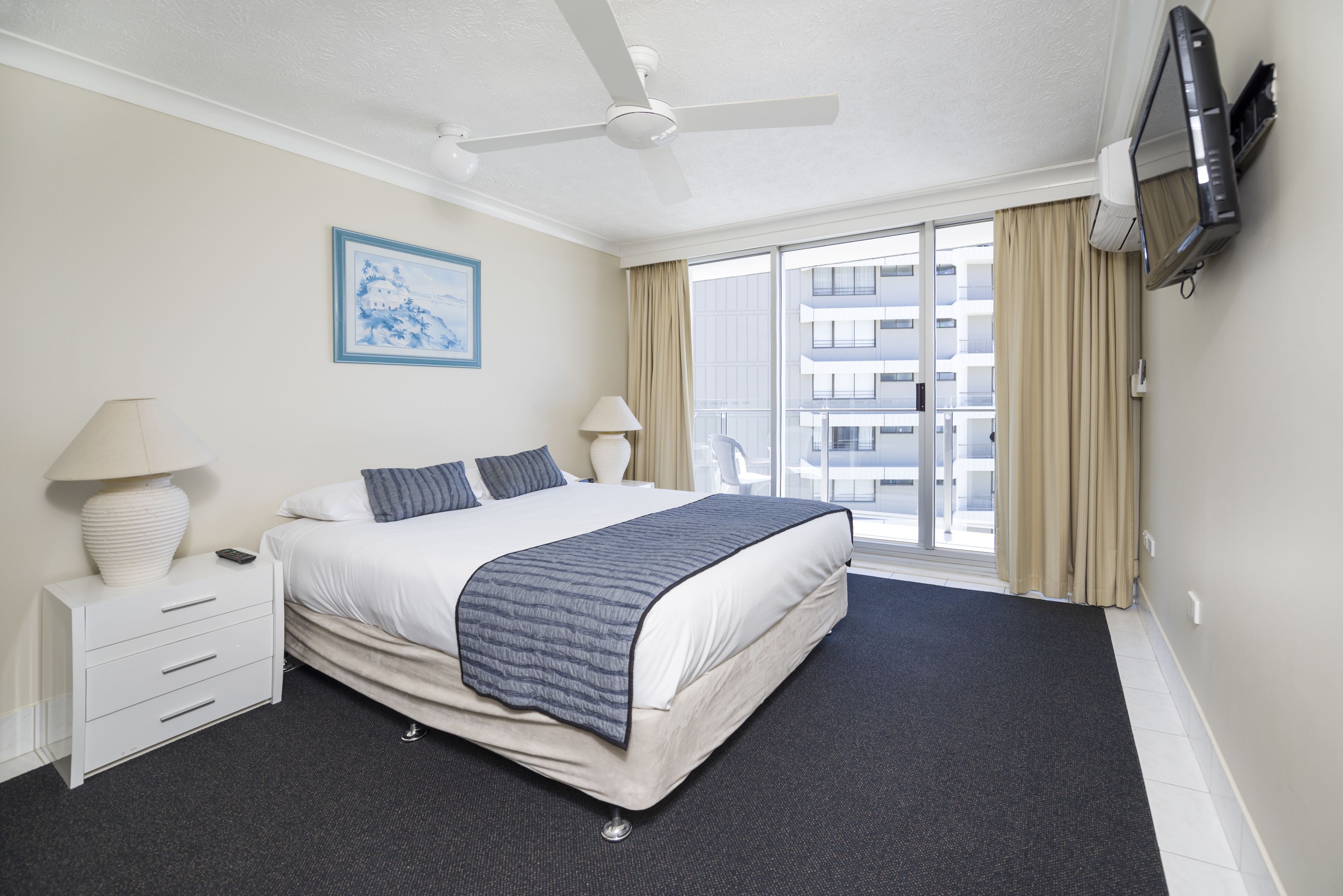 A hotel room with a bed, a ceiling fan, a TV, a window with curtains, and a painting on the wall.