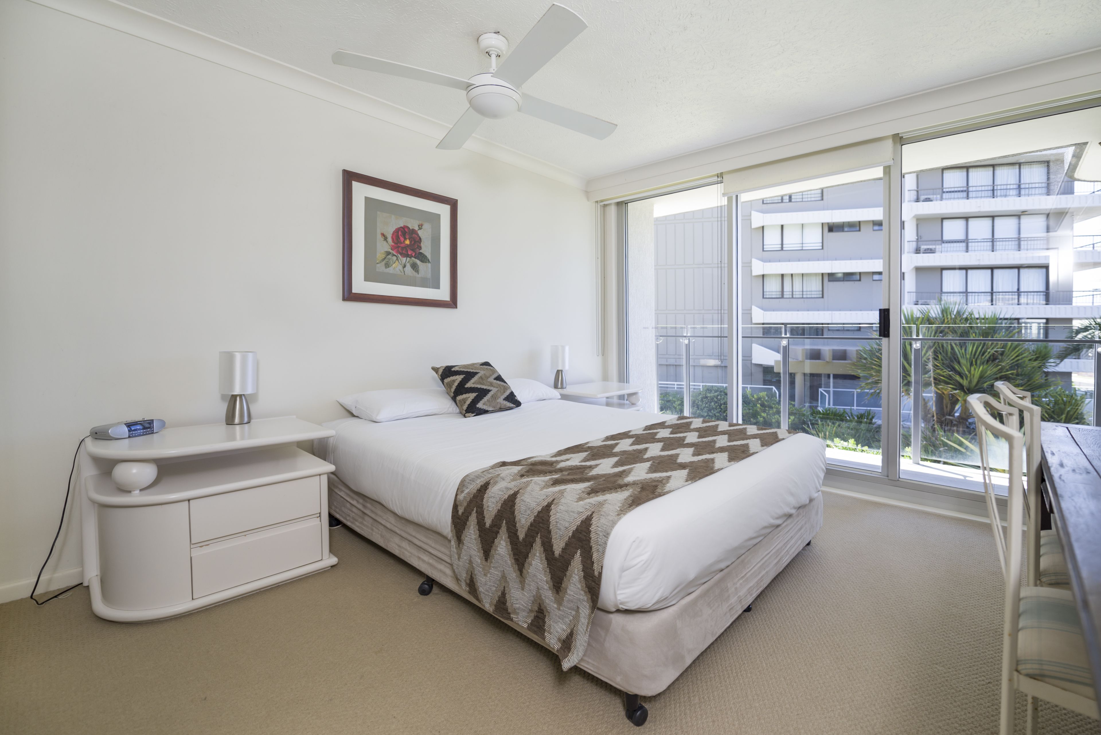 A bedroom with a bed, nightstand, bedside table, ceiling fan, and a large window with a view of buildings.