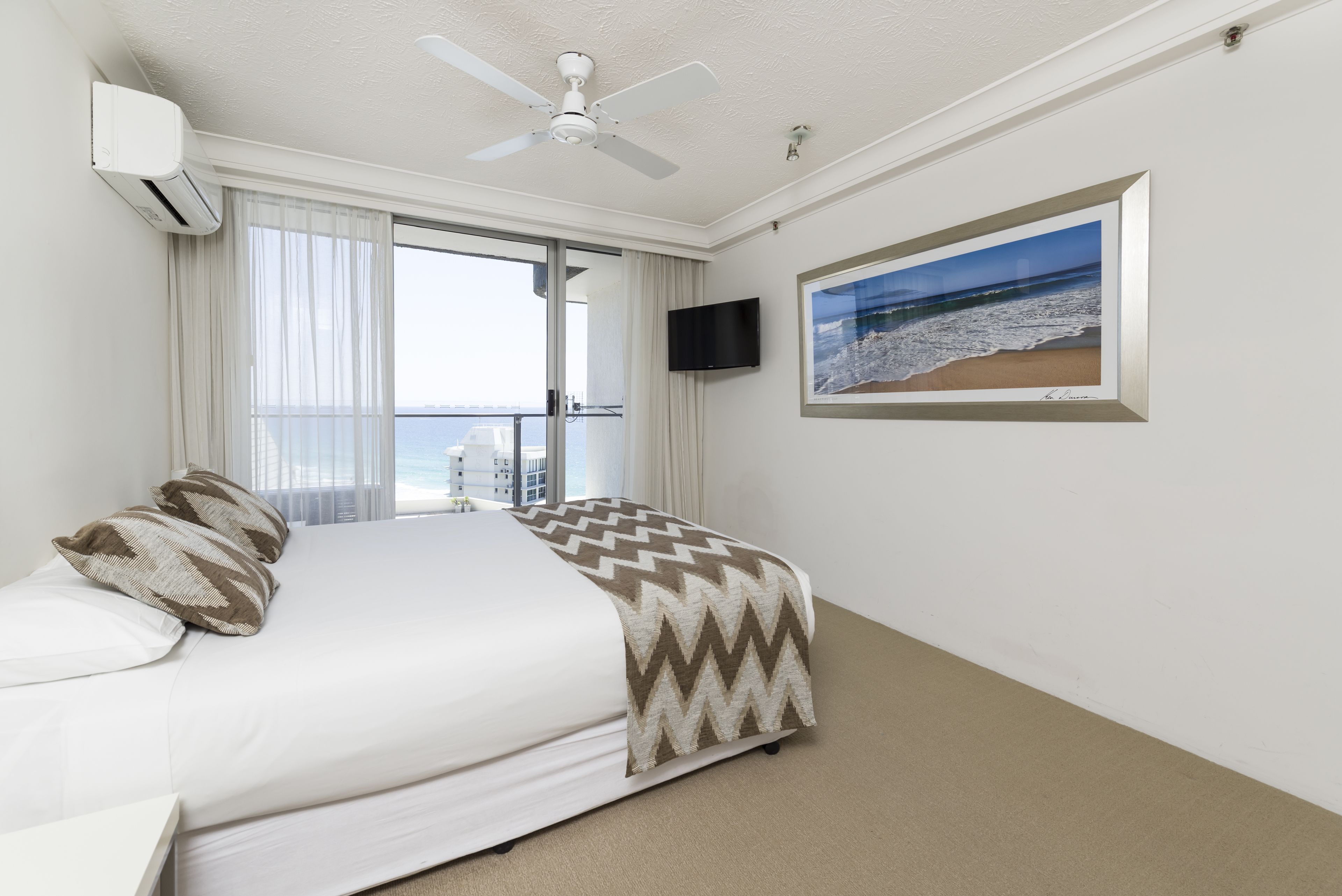 Une chambre d’hôtel avec un lit, un ventilateur de plafond, une télévision et une grande fenêtre donnant sur la ville.