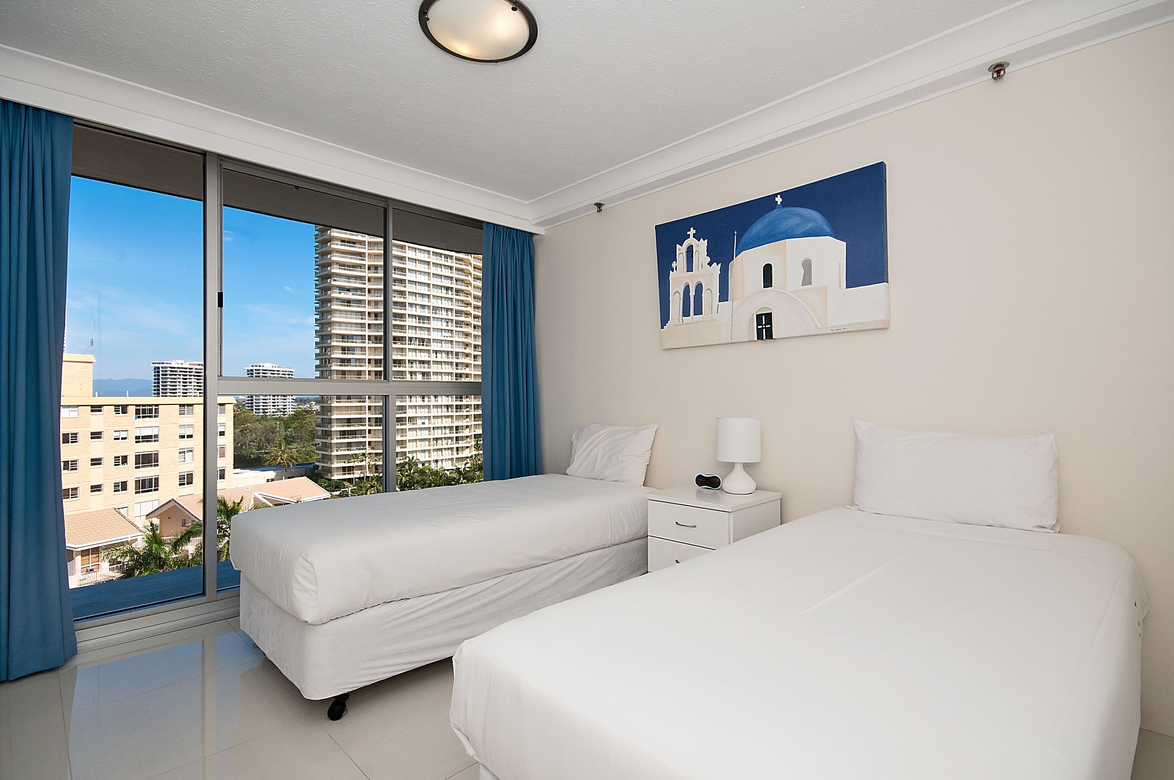 A hotel room with two beds, a nightstand, a lamp, and a painting of a building.