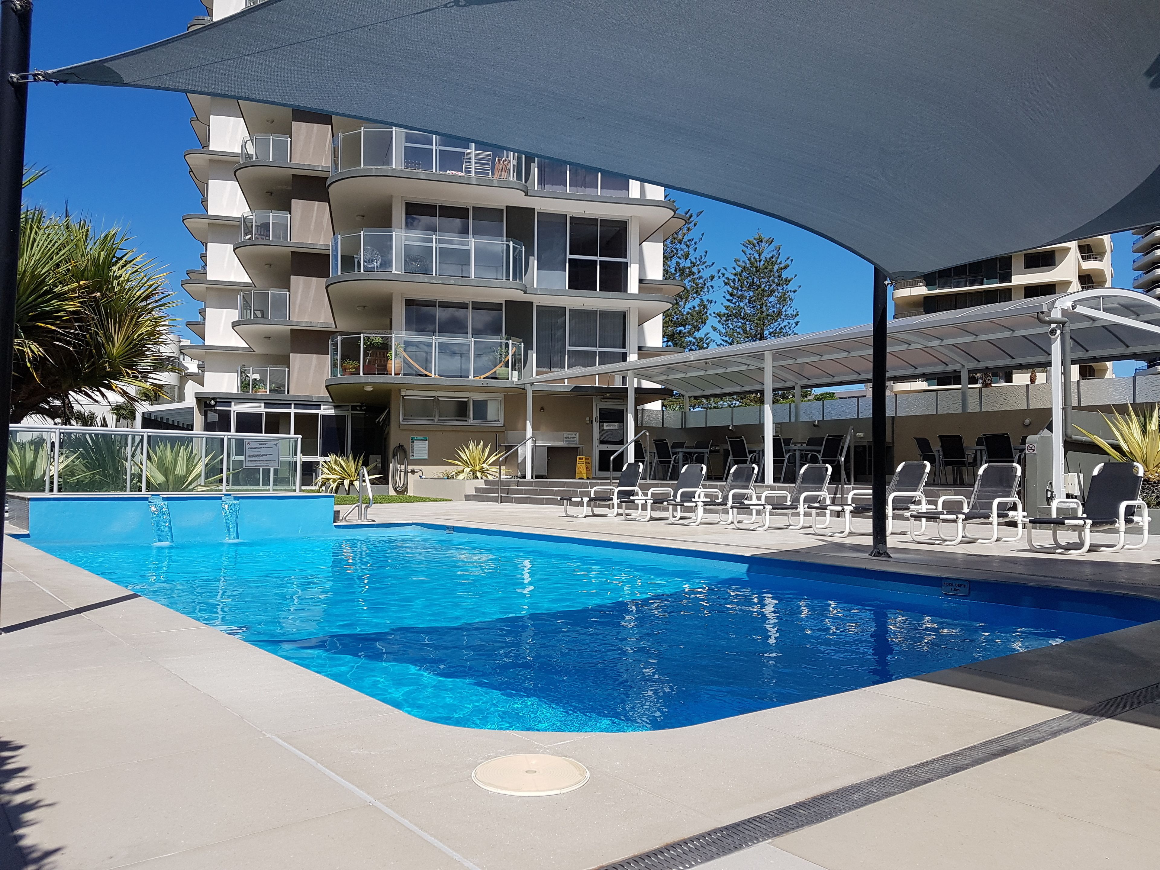 A swimming pool with lounge chairs and a covered seating area, surrounded by a multi-story building with balconies.