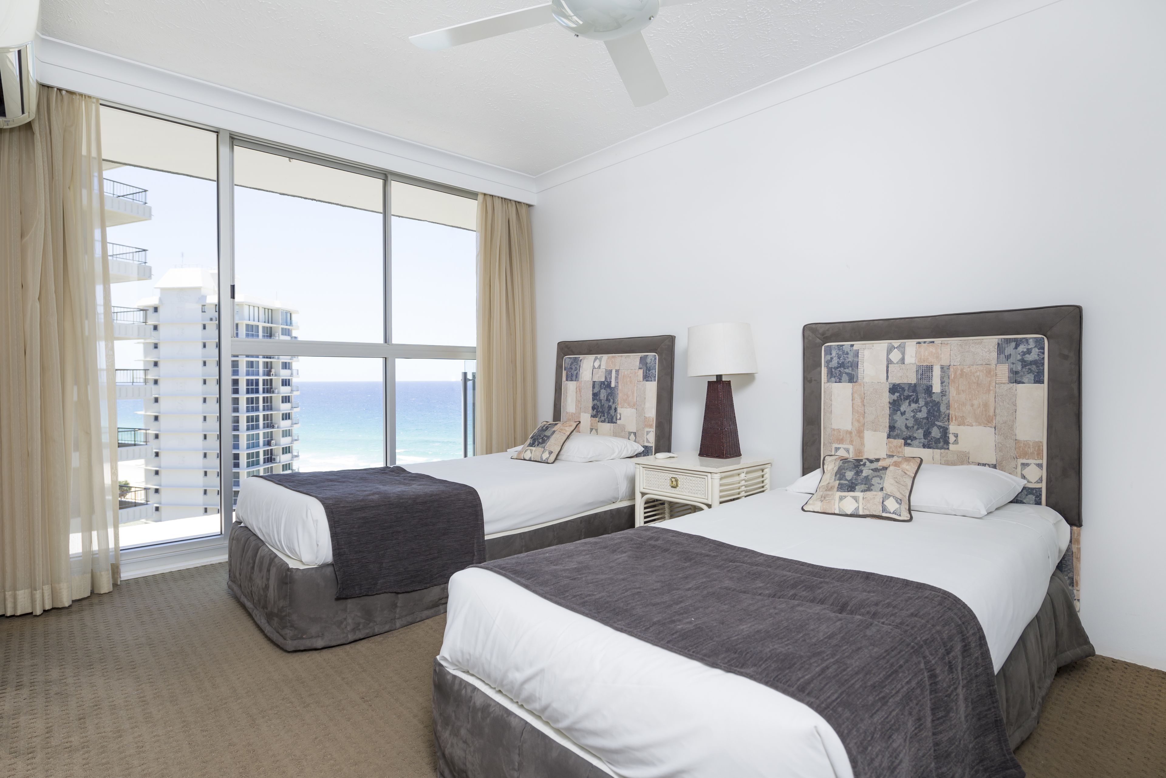 A hotel room with two beds, a large window with a view of the ocean, and a ceiling fan.