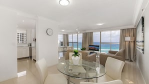 A modern living room with a glass table, white furniture, a view of the ocean, and a dining area with a kitchen in the background. - Sunbird Beach Resort (Main Beach)