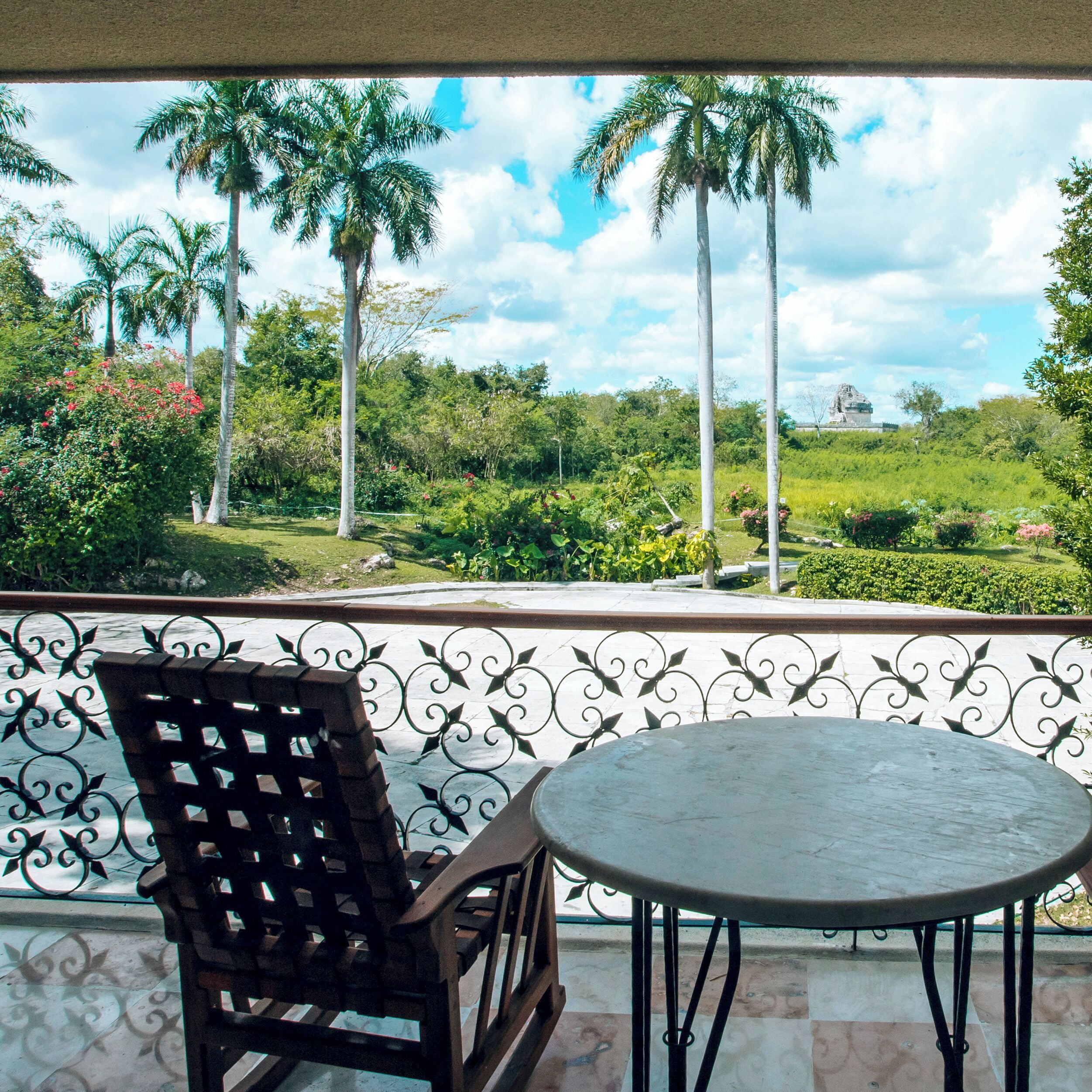 A balcony with a view of palm trees and a statue, a wooden chair, and a round table.