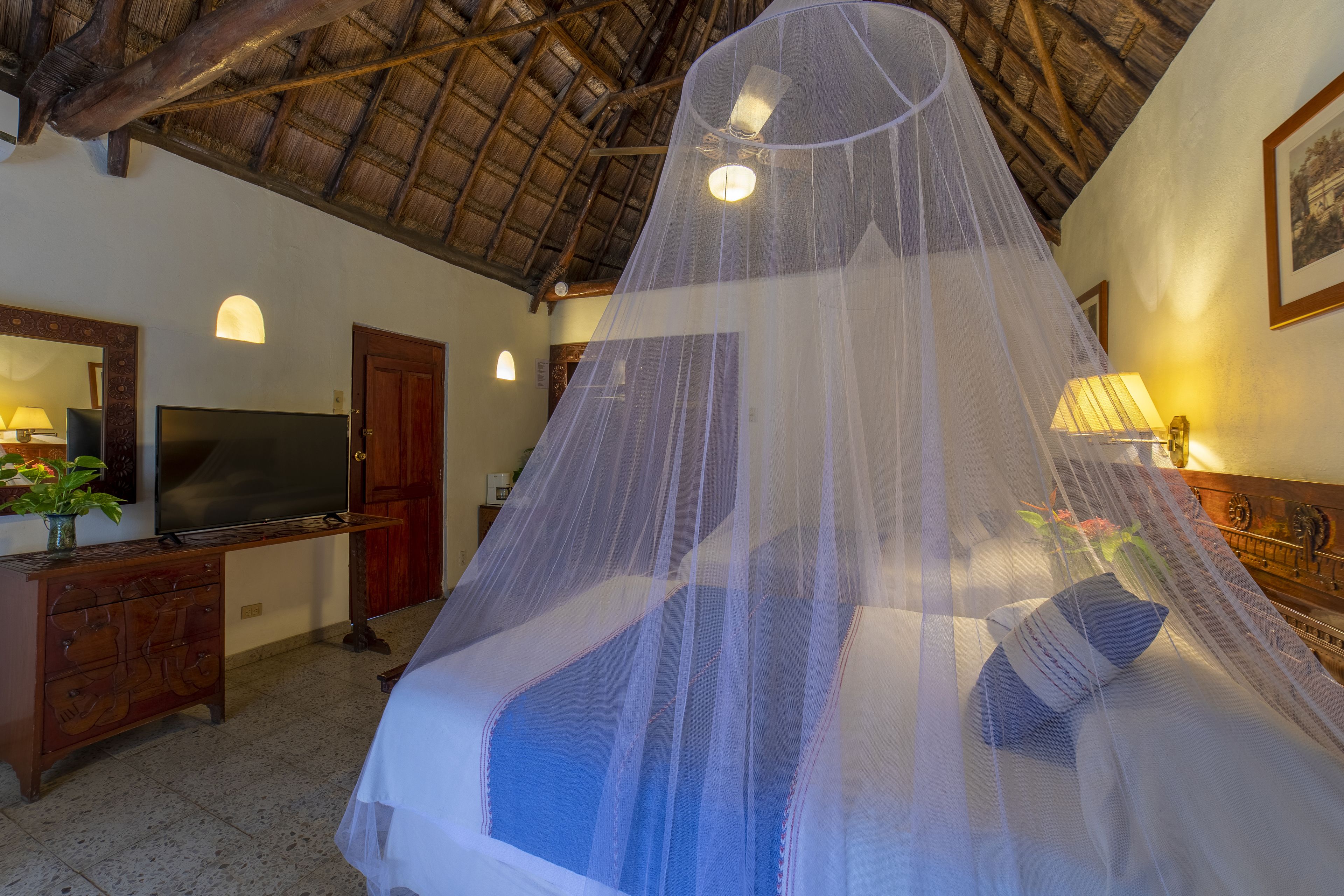 Una habitación con una cama bajo una mosquitera, un televisor, un tocador de madera y un escritorio con una lámpara.
