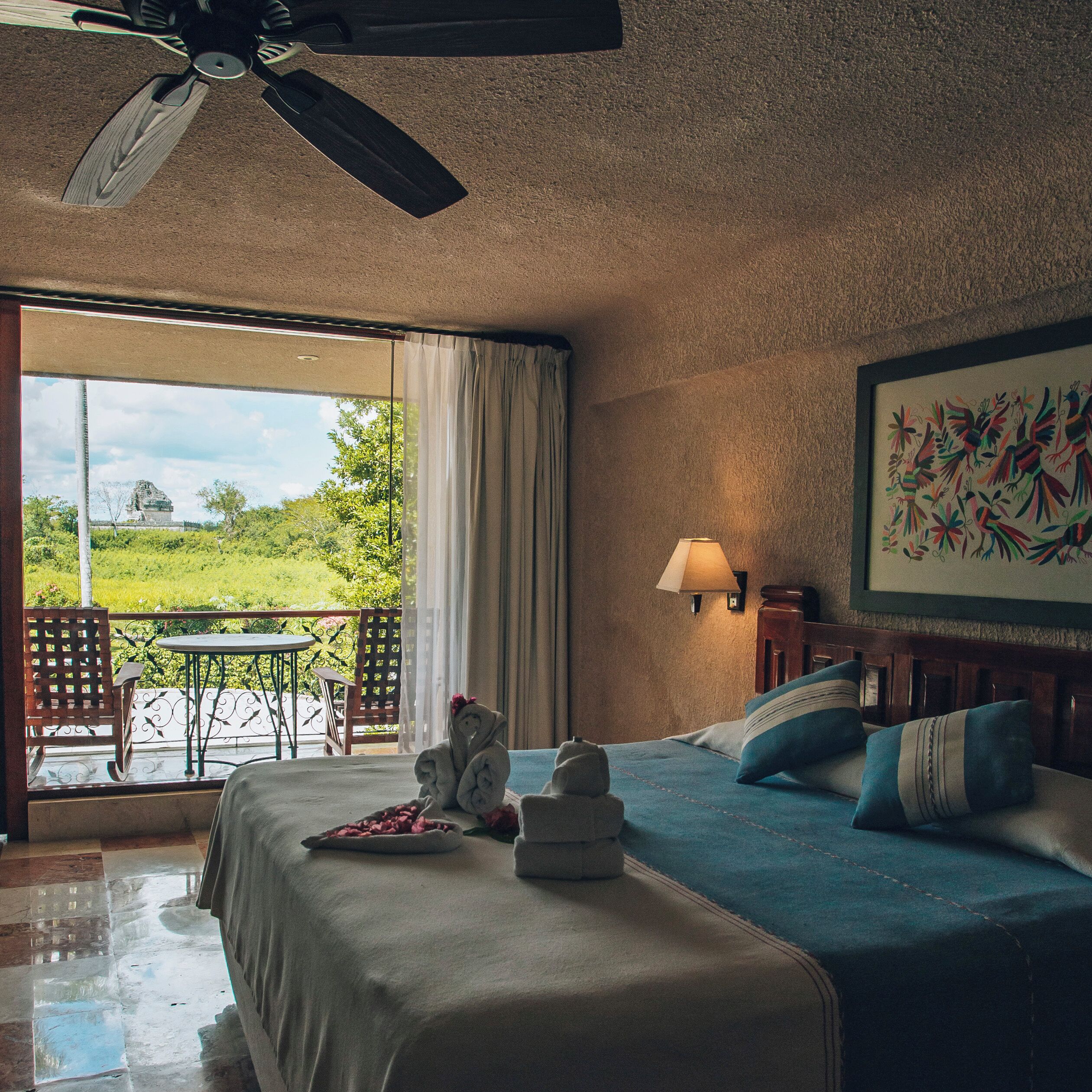 A hotel room with a bed, a table, a chair, a ceiling fan, and a view of the outdoors.