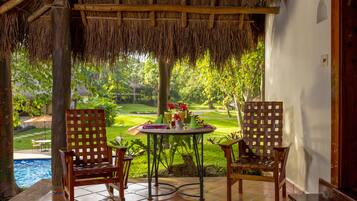 A patio with a thatched roof, two wooden chairs, a round table with a drink and a flower, and a view of a pool and garden.