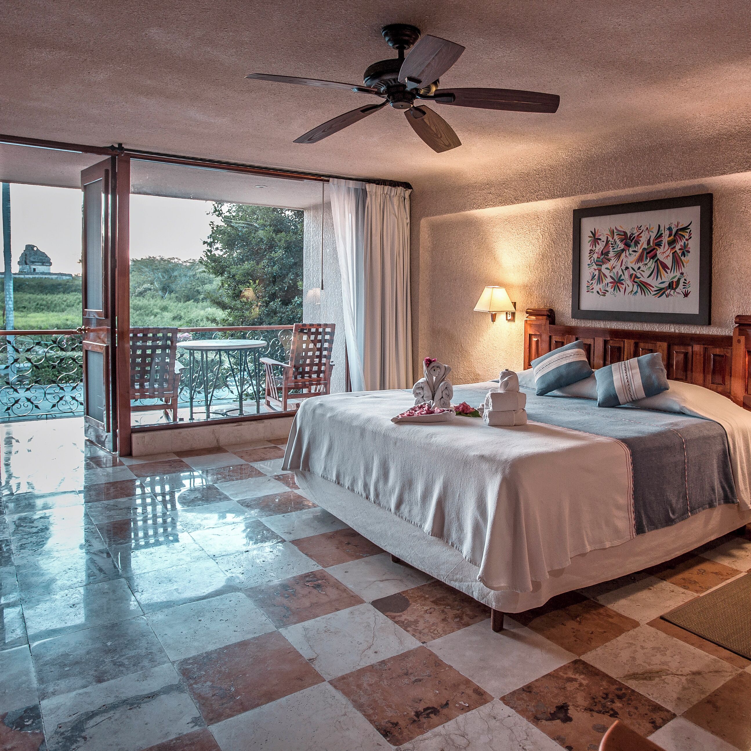 A hotel room with a bed, a balcony, a ceiling fan, and a painting on the wall.