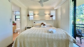 Quarto com cama, ventoinha no teto, mesa de cabeceira e janela com persianas.