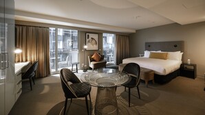 A hotel room with a large bed, a desk with a chair, a round glass table, and a view of the city through large windows. - The Kinson (Melbourne)