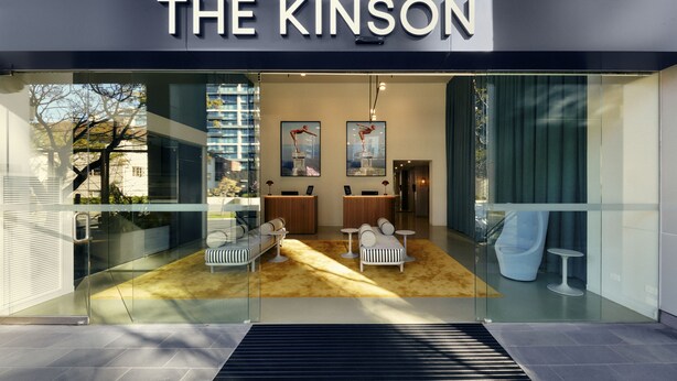 A modern hotel entrance with a glass facade, a reception desk, seating area, and a view of the cityscape. - The Kinson (Melbourne)