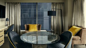 A modern hotel room with a round glass table, two chairs, a sofa with yellow cushions, a TV, and a large window with curtains. - The Kinson (Melbourne)