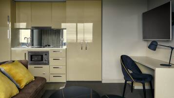 Een ruimtelijk ingerichte woonruimte met kitchenette, een bank met gele kussens, een bureau met stoel en een flatscreen televisie aan de muur.